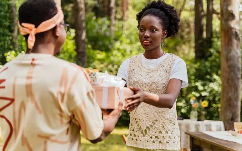 A woman hands a gift to another person during an outdoor celebration in a forest setting.