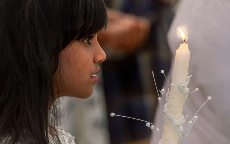 A young girl focuses intently while holding a beautifully decorated candle during a solemn indoor ceremony.