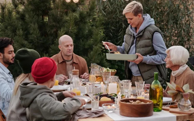 Smiling woman wearing warm vest and hoodie serving baked dish during holiday party at big table under garlands and evergreen trees beside