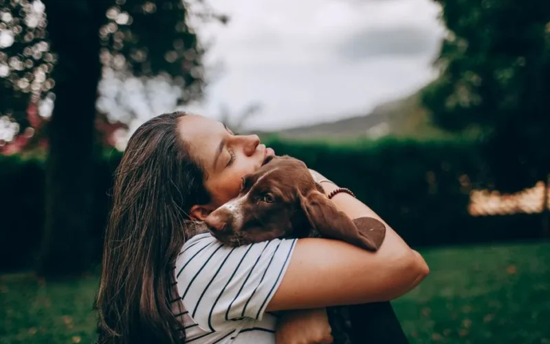 A woman lovingly embraces her dog in a green outdoor space, highlighting companionship.
