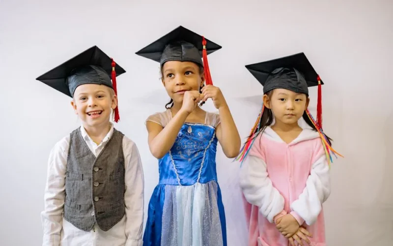 Three children in graduation caps express pride and joy at their preschool graduation ceremony.