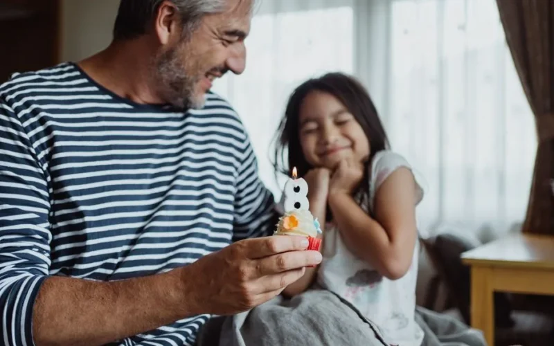 A joyful father and daughter duo celebrating her 8th birthday indoors with a cupcake.
