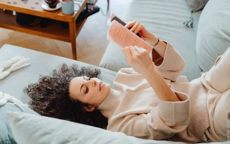 Woman in sweater reading a book on a cozy sofa, enjoying leisure time indoors with a cup of tea.