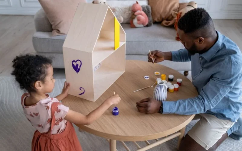 A father and daughter enjoy painting a wooden dollhouse together indoors.