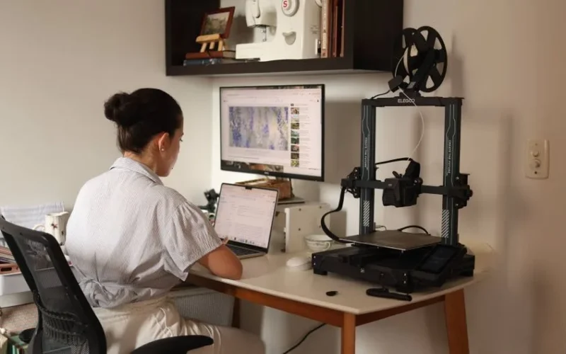 A woman working on a laptop with a 3D printer in a modern home office setup.