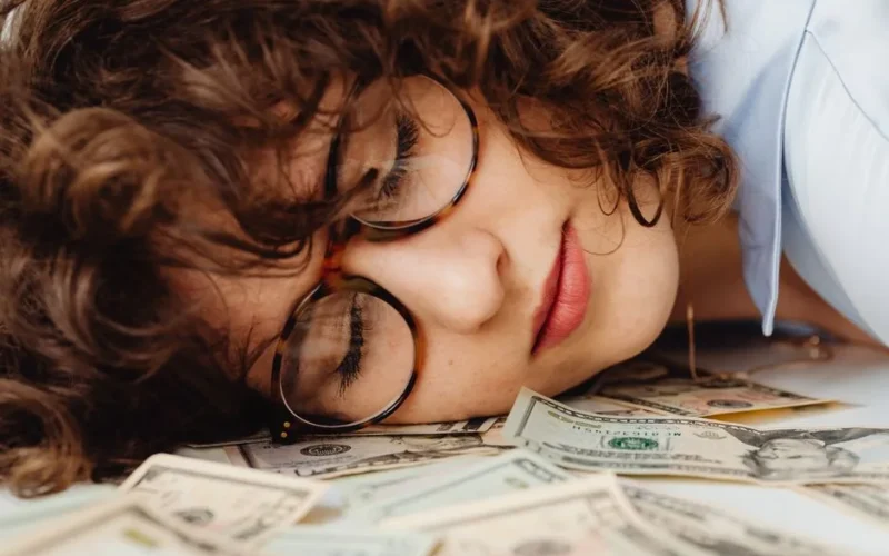 Close-up of a sleeping woman with curly hair and eyeglasses, resting on a pile of cash.