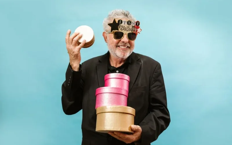 Bearded senior man in festive sunglasses holding birthday gifts against a blue background.