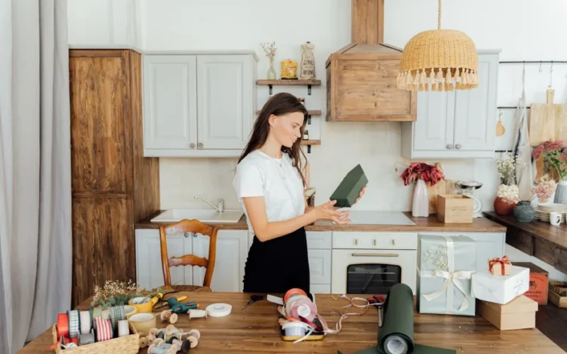 A woman wrapping presents in a cozy kitchen with ribbon and paper around.