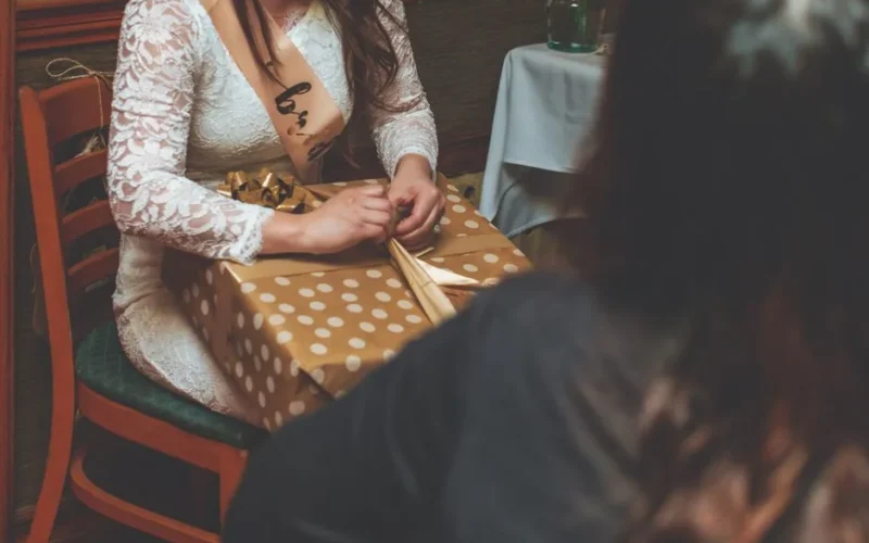 A woman at a bridal shower unwrapping a polka dot gift indoors, wearing a sash.