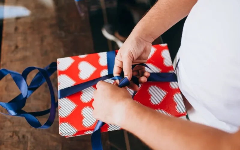 Hands tying a blue ribbon on a heart-patterned gift, symbolizing love and celebration.