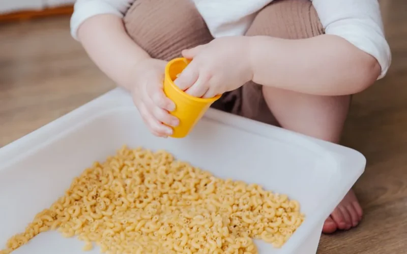 A young child plays with dry macaroni in a sensory activity, enhancing creativity and learning.