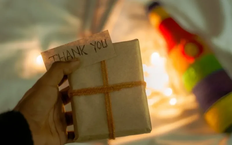 A close-up of a hand holding a gift with a 'Thank You' note, wrapped in brown paper and twine, with warm lights.