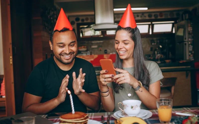 Two young adults celebrating a birthday with cake and coffee indoors, wearing party hats.