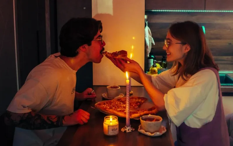 Couple shares a romantic candlelit pizza dinner in a cozy kitchen setting.