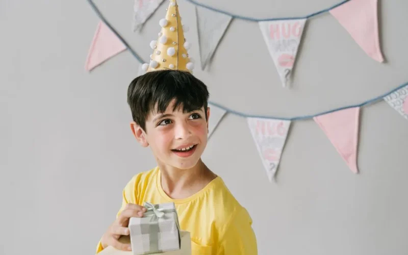 Young boy wearing party hat and holding a gift, celebrating birthday indoors.