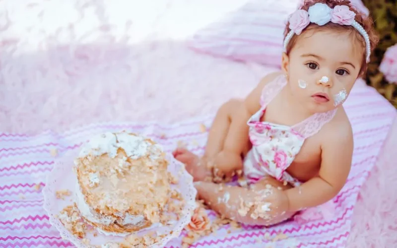 Cute baby enjoying a cake smash for her first birthday, sitting outdoors on a blanket.