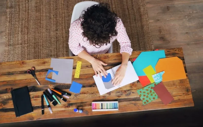 Person engaging in artistic crafts with colorful paper and tools on a wooden table.