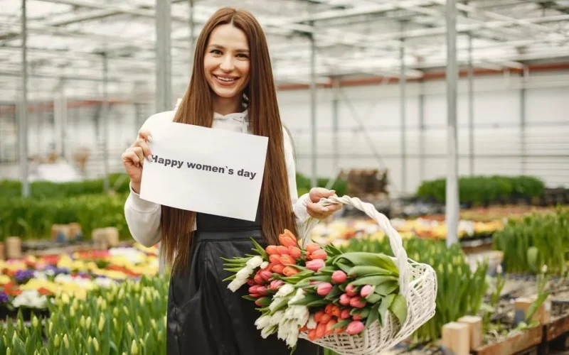 Woman with long hair smiling, holding flowers and a sign for women's day in a vibrant greenhouse.