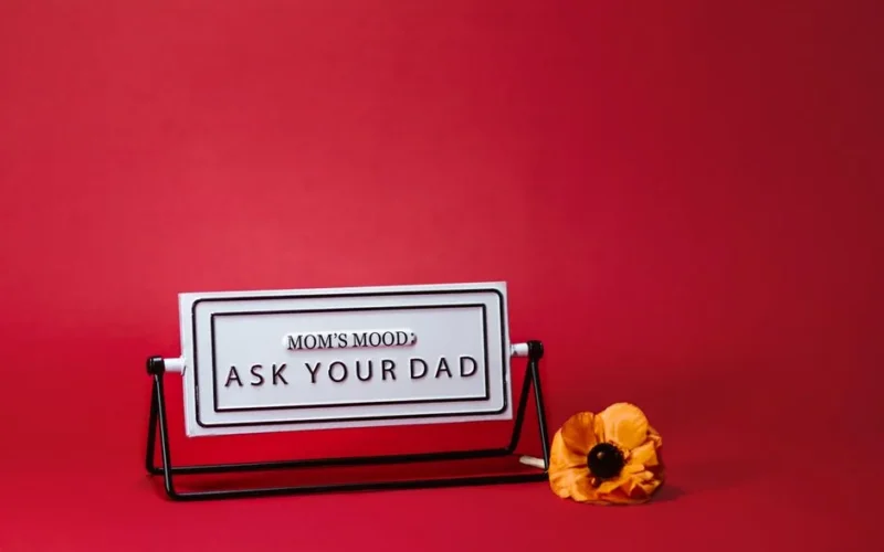 A whimsical Mother's Day sign with 'Mom's Mood: Ask Your Dad' text, set against a red backdrop with a flower.