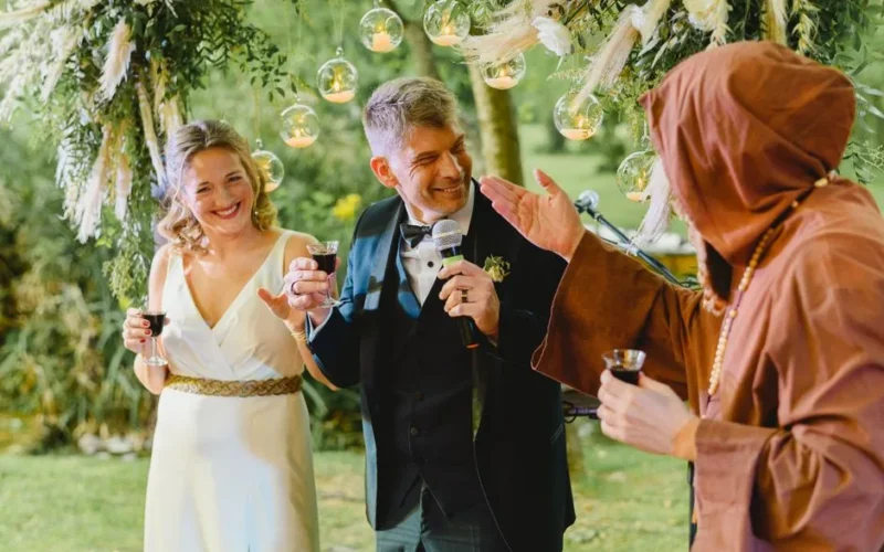 Bride and groom share a joyful toast with monk at outdoor wedding ceremony decorated with greenery and hanging lights.
