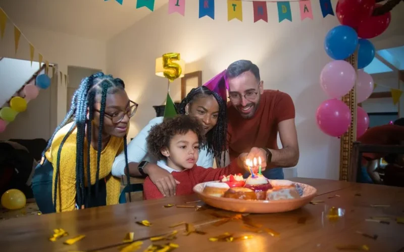 Joyful family celebrating a child's 5th birthday with cake and candles indoors.