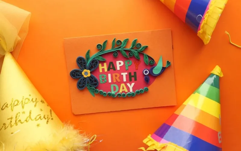 Colorful quilling birthday card surrounded by party hats on an orange backdrop, perfect for festive celebrations.