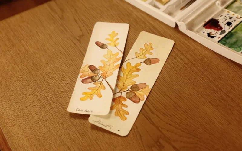 Artistic bookmarks featuring watercolor oak leaves and acorns on a wooden table.