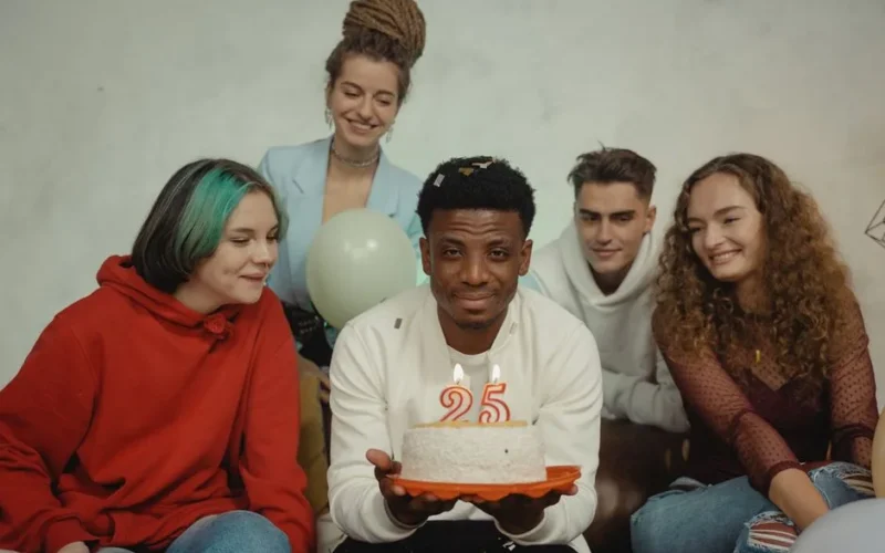 A joyful group of friends celebrating a 25th birthday indoors with a cake and candles.