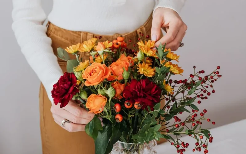 A woman arranging a colorful floral bouquet with roses and chrysanthemums in a glass vase.