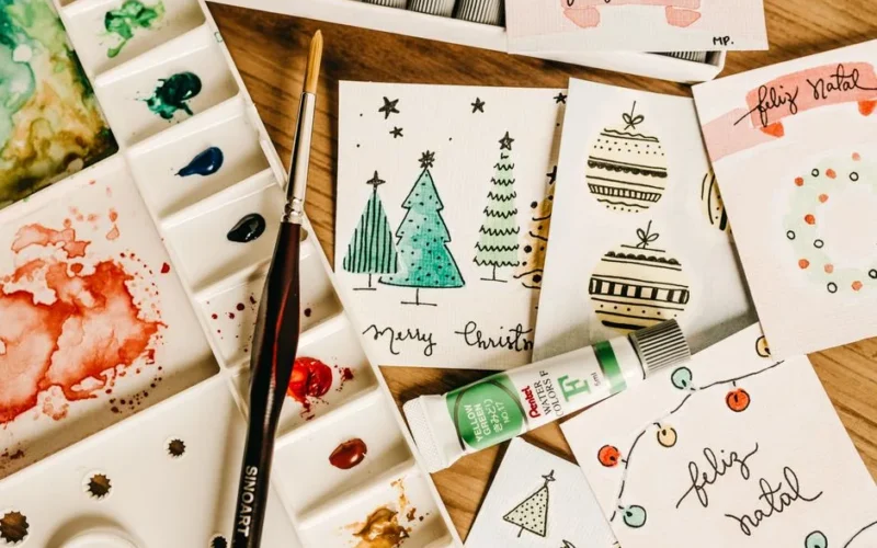 Artistic holiday card designs with watercolor paints and brushes on a wooden table.