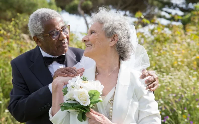 Smiling elderly couple in wedding attire embracing outdoors with flowers.