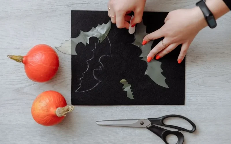 Hands crafting Halloween bat decorations on black paper with scissors and pumpkins.