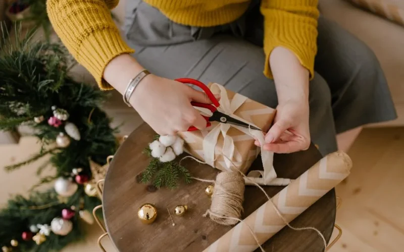 An adult wrapping a Christmas gift with ribbons and decorations at home.