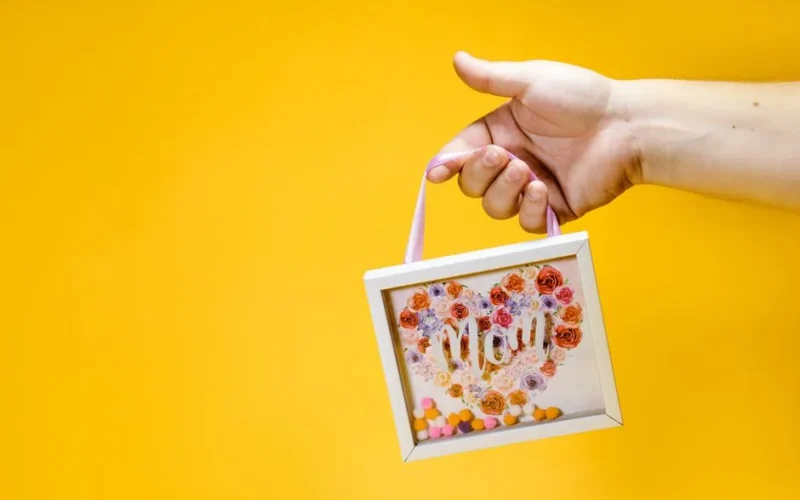 Close-up of a hand holding a floral-themed Mother's Day gift on a vibrant yellow background.