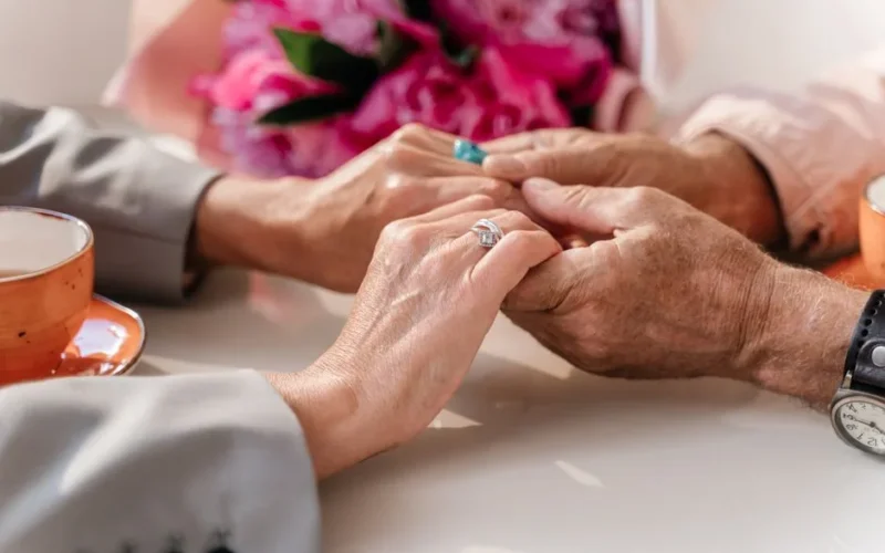 An elderly couple shares a tender moment, holding hands over a table with coffee and flowers.