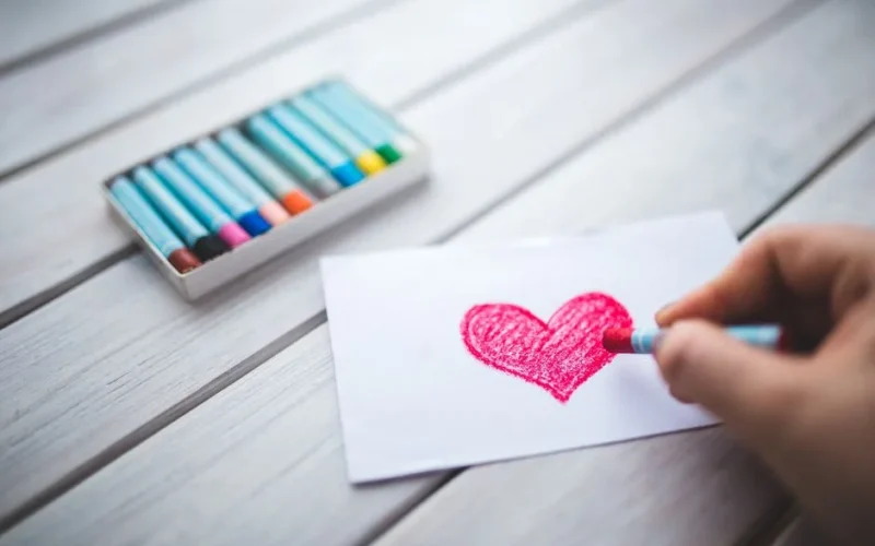 A close-up of a hand using pastel crayons to draw a red heart on paper.