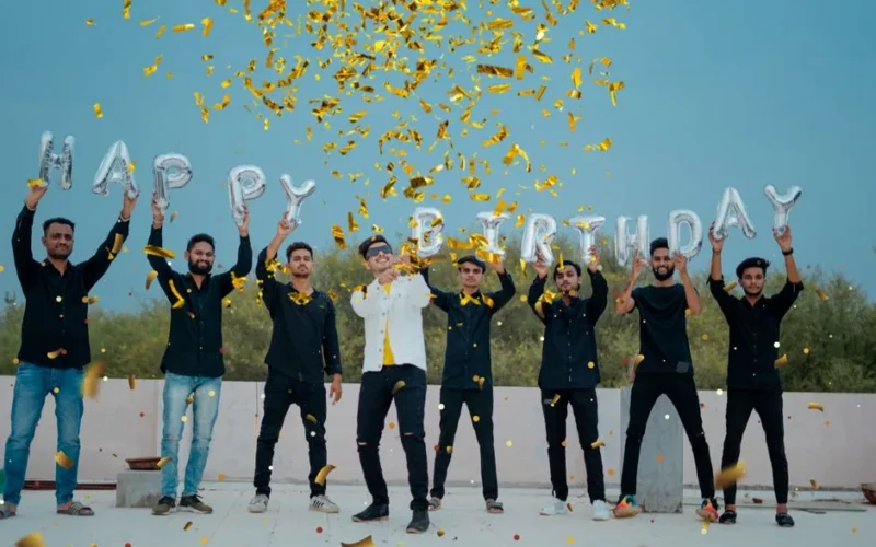 A joyful group of young men celebrates a birthday outdoors with confetti and balloon letters.