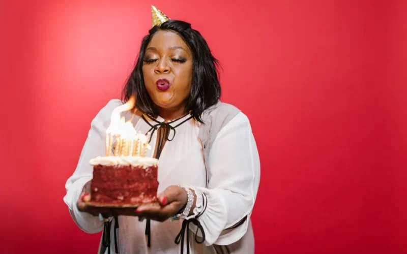 A black adult woman joyfully blowing out birthday candles on a red velvet cake against a red backdrop.