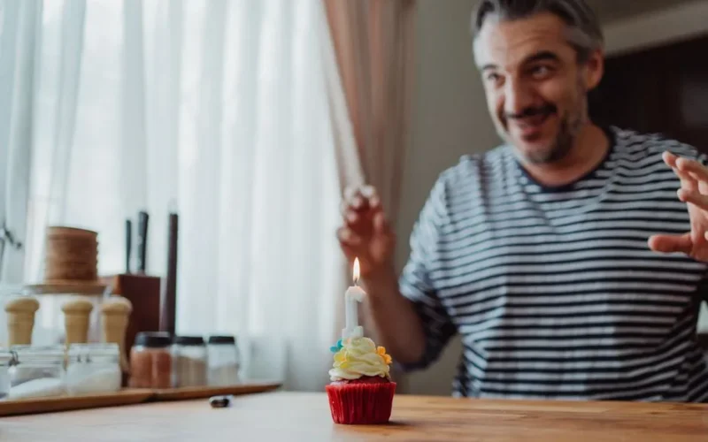 A joyful man in a striped shirt celebrates with a cupcake topped with a lit candle.