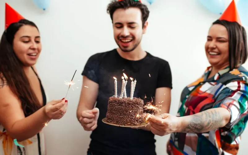 Three friends celebrate a birthday with cake and sparklers indoors wearing party hats.