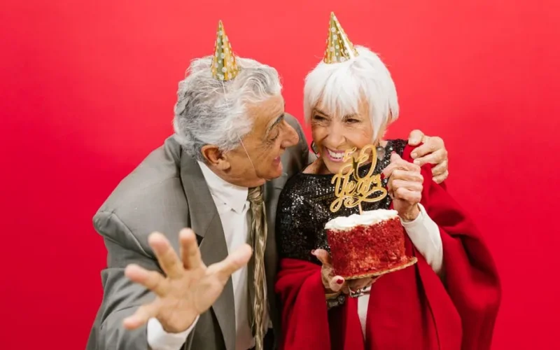 Elderly couple with party hats celebrating 50th anniversary with a cake and laughter.