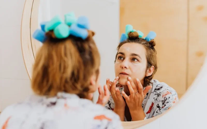 Woman with curlers applying cream in front of a mirror indoors.
