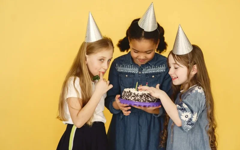 Three children enjoying a birthday party with cake and party hats on a yellow background.