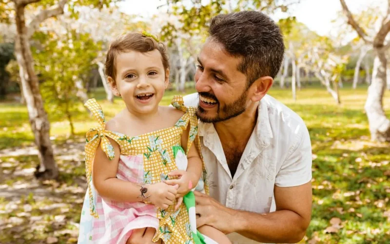 A joyful father and daughter share a moment of laughter in a sunlit orchard setting.