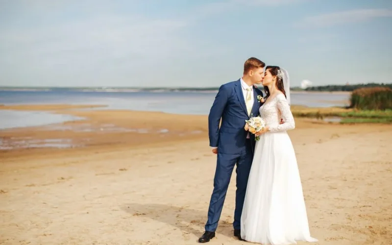 Bride and groom sharing a kiss on a beautiful sandy beach under a clear blue sky.
