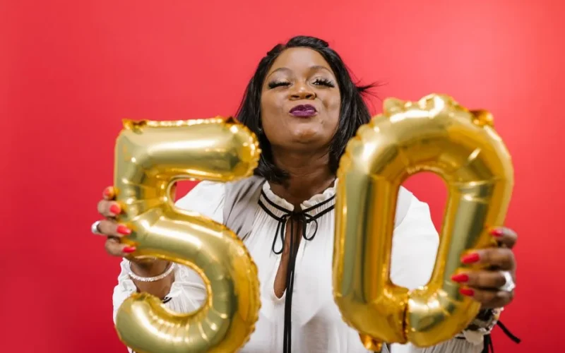Joyful woman holding golden 50 balloons against a vibrant red background.