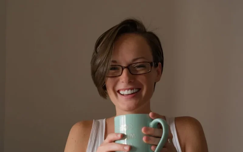 Happy woman wearing glasses and holding a mug, smiling warmly inside a cozy room.