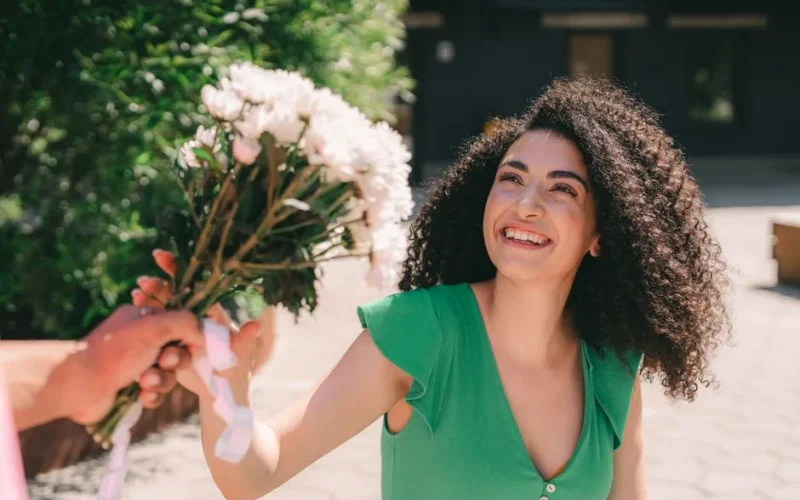 A woman smiles brightly while receiving a bouquet in an outdoor urban setting.
