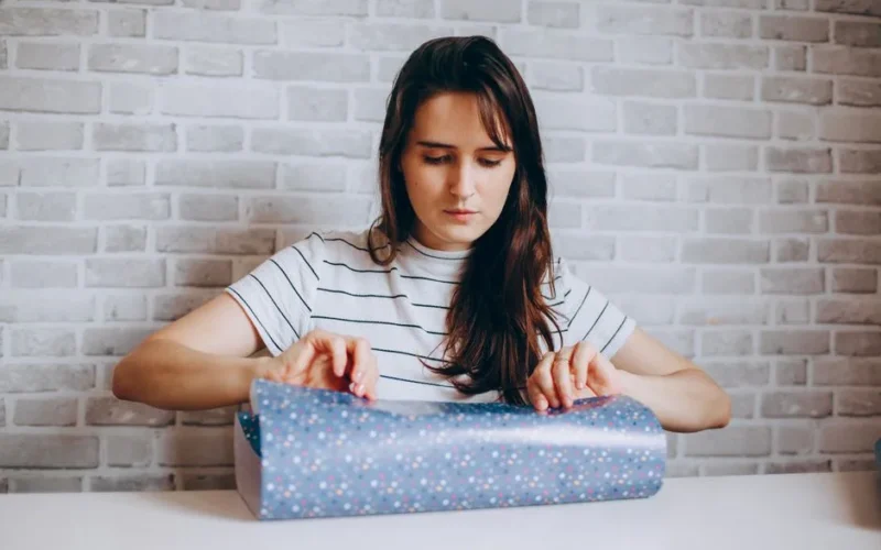 Young woman focused on wrapping a gift against a stylish brick wall indoors.