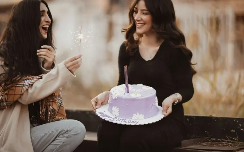Two women celebrating a birthday outdoors, smiling with a purple cake and sparklers.
